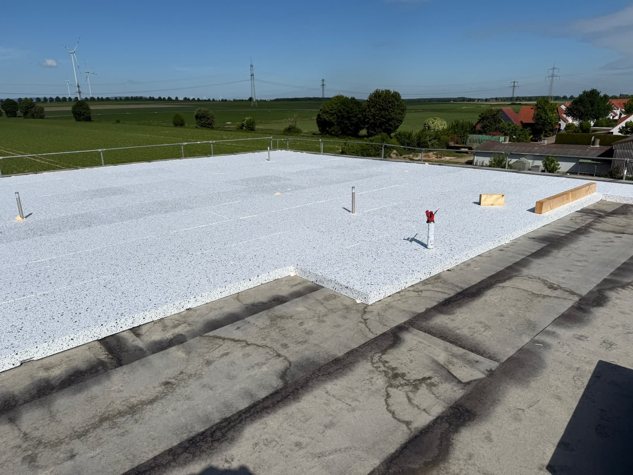 Ein im Bau befindliches Flachdach mit installierten Dämmplatten. Mehrere Rohre ragen aus der Oberfläche heraus, und in der Nähe des Randes sind Holzblöcke angebracht. Der Hintergrund zeigt Felder, Bäume und entfernte Häuser unter einem klaren Himmel.
