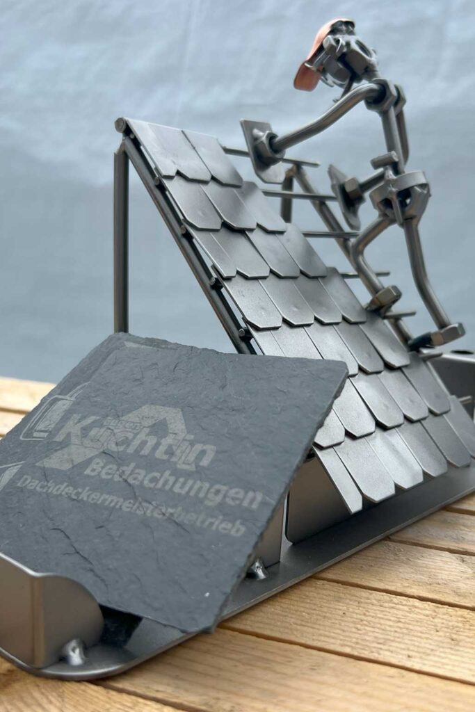 Eine Metallskulptur zeigt einen Dachdecker, der Schindeln auf einem Schrägdach befestigt. Die Skulptur steht auf einer Holzfläche. Im Vordergrund ist eine Schieferplatte mit aufgedrucktem Text zu sehen.