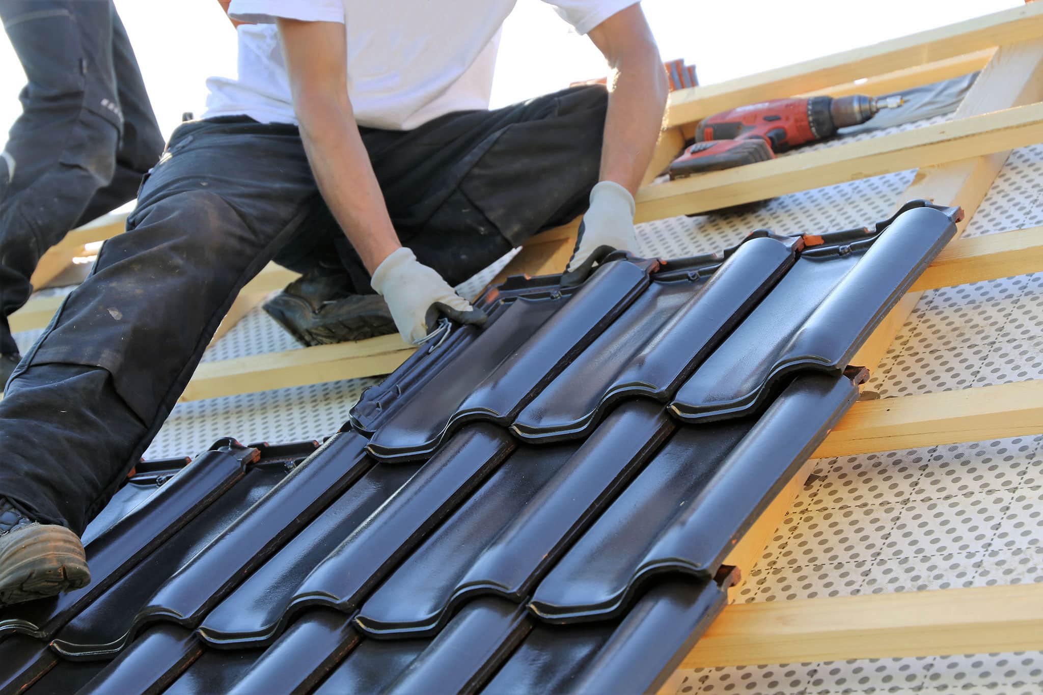 Eine Person mit Handschuhen und Arbeitskleidung verlegt schwarze Keramikdachziegel auf einem Holzgerüst, wobei eine Bohrmaschine in der Nähe zu sehen ist.