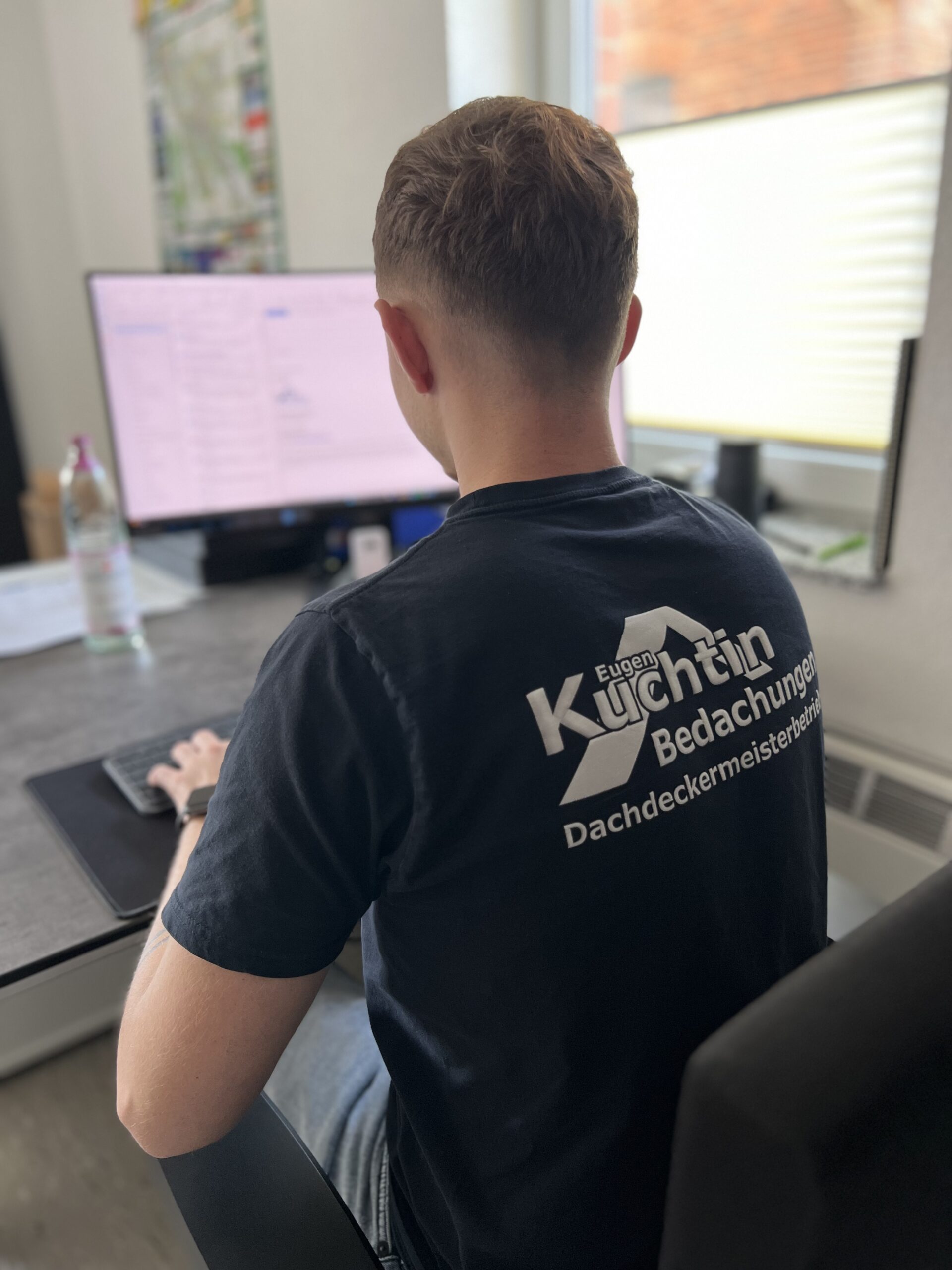 Eine Person mit kurzen Haaren sitzt an einem Schreibtisch und tippt auf einer Tastatur vor einem Monitor. Auf der Rückseite ihres Hemdes steht „Eugen Küchtin Bedachungen Dachdeckermeisterbetrieb“. Auf dem Schreibtisch steht eine Wasserflasche.