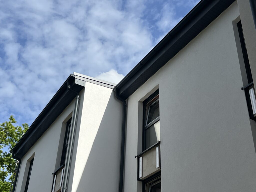Das Bild zeigt den oberen Teil eines modernen weißen Gebäudes mit dunklen Fensterrahmen und Regenrinnen vor einem teilweise bewölkten Himmel. Auf der linken Seite sind einige Äste zu sehen.