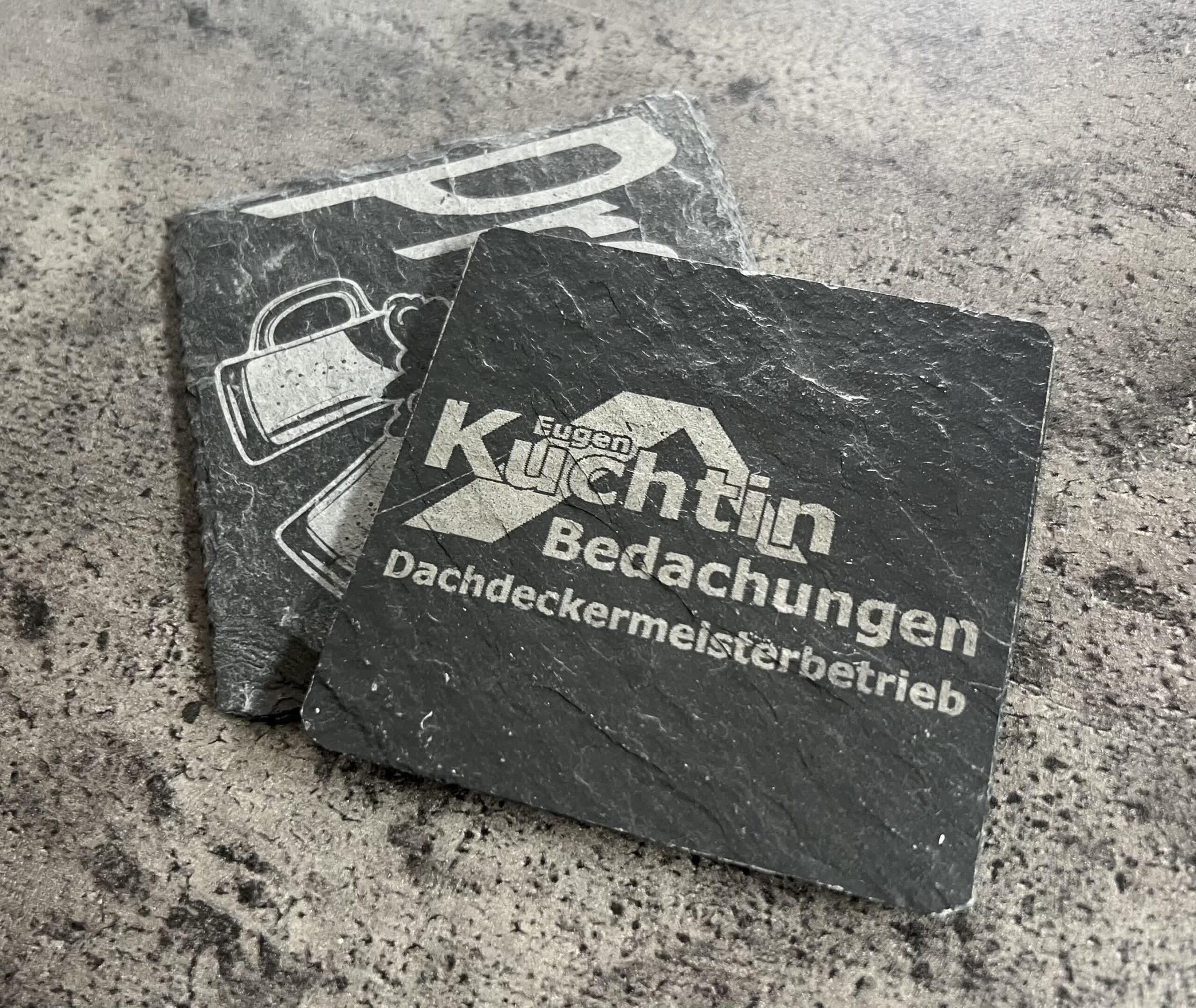 Zwei quadratische Schieferuntersetzer sind auf einer strukturierten Oberfläche abgebildet. Der vordere Untersetzer zeigt den Text "Kuchtn Bedachungen Dachdeckermeisterbetrieb", während der hintere Untersetzer einen Teil einer Grafik und einen Teil des Textes zeigt.