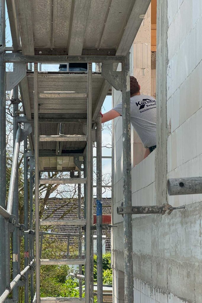 Eine Person in einem grauen T-Shirt arbeitet an der Außenwand eines im Bau befindlichen Gebäudes. Neben dem Gebäude ist ein Metallgerüst aufgebaut, im Hintergrund sind Bäume und Dächer zu sehen.