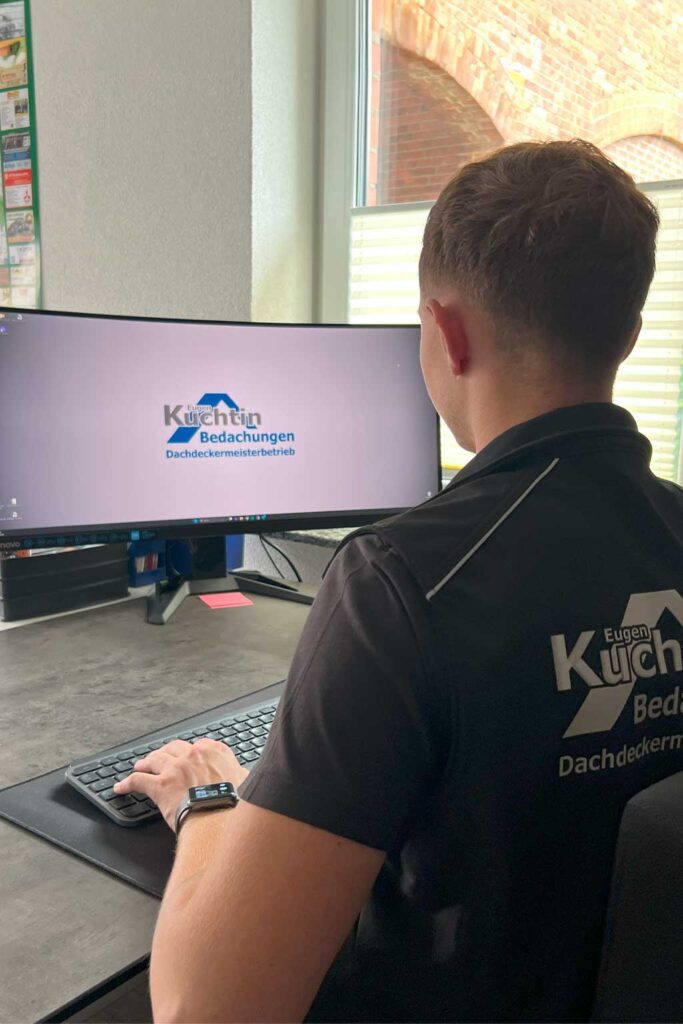 Ein Mann, der ein schwarzes Hemd mit dem Logo „Kuchen Bedachungen Dachdeckermeisterbetrieb“ trägt, arbeitet in einer Büroumgebung an einem Computer mit einem gebogenen Monitor, auf dem dasselbe Firmenlogo angezeigt wird.