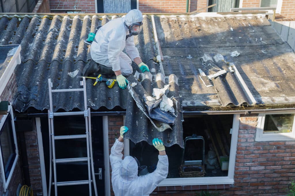 Zwei Personen in Schutzanzügen und mit Masken entfernen mit Werkzeugen Wellasbestdachplatten von einem Gebäude. Eine Person steht auf dem Dach, während die andere von unten in der Nähe einer Leiter assistiert.