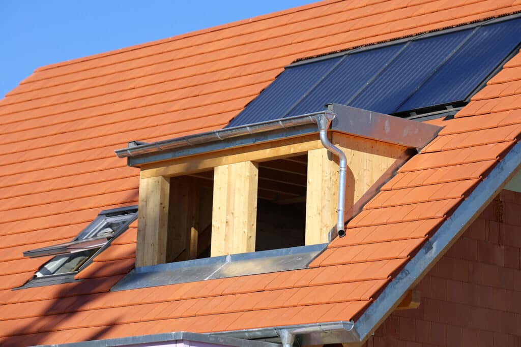 Nahaufnahme eines Hausdachs mit orangefarbenen Ziegeln, das eine teilweise ausgebaute Dachgaube, Metalldachrinnen und auf dem Dach installierte Solarzellen zeigt.