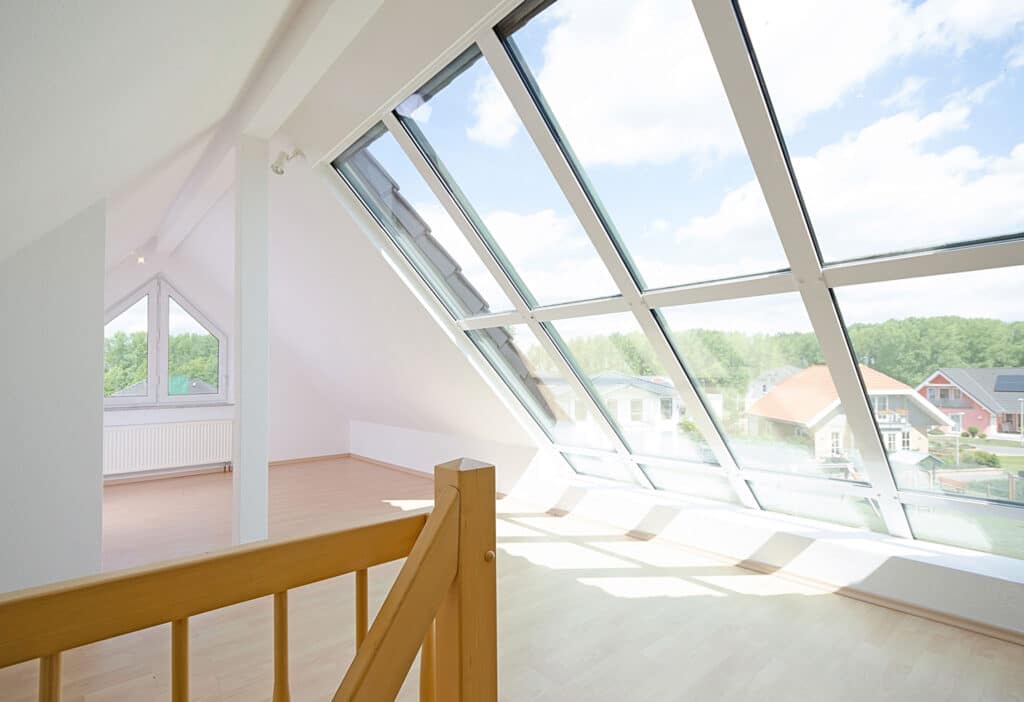 Ein heller, leerer Dachraum mit schrägen Glasfenstern an einer Wand, hellem Holzfußboden, weißen Wänden und einem Holzgeländer, das von einer Treppe nach oben führt. Durch die Fenster sind draußen Häuser und Bäume zu sehen.