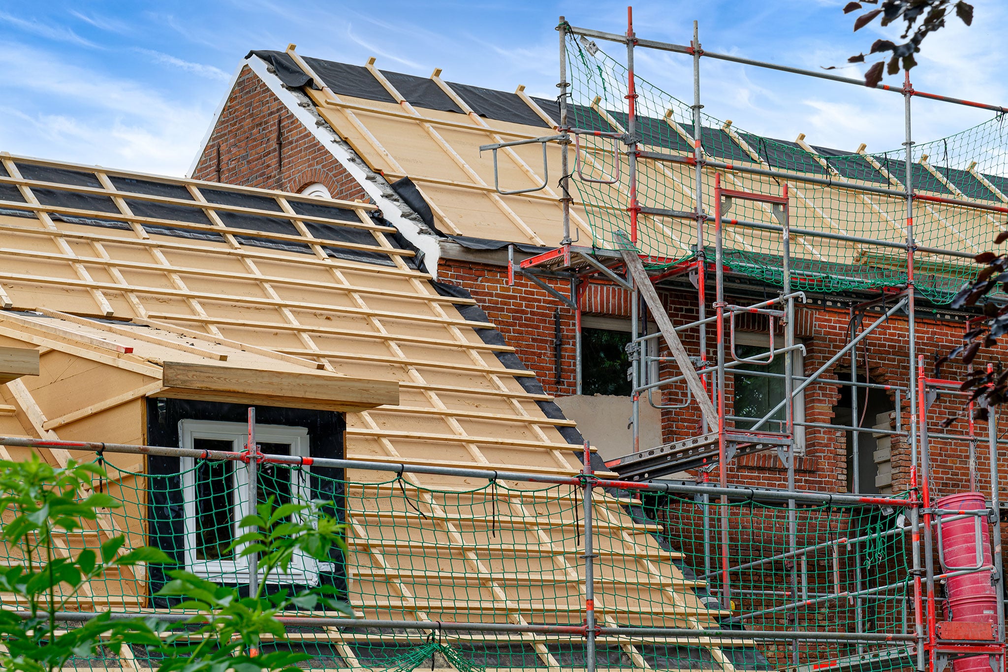 Ein im Bau befindliches Backsteinhaus mit hölzernem Dachstuhl und Gerüst. Das Dach ist teilweise mit Holzbalken bedeckt, und ein Sicherheitsnetz umgibt das Gerüst. Der Himmel ist blau und leicht bewölkt.
