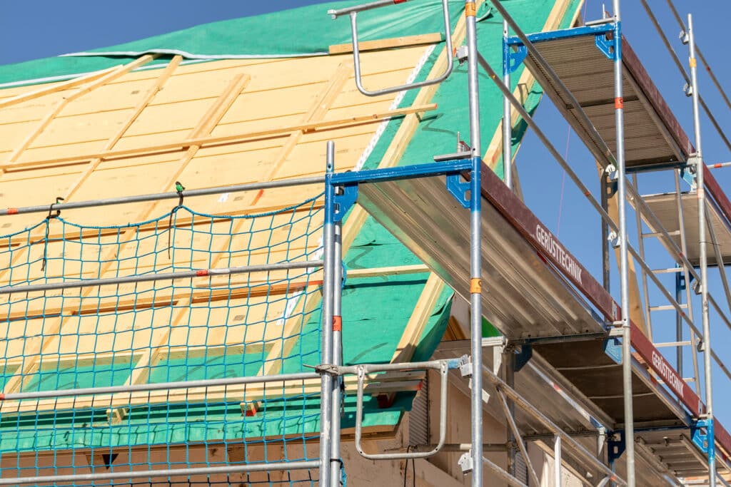 Ein teilweise fertiggestelltes Gebäudedach, bedeckt mit grüner Folie und Holzpaneelen und umgeben von einem Metallgerüst unter einem klaren blauen Himmel. Auf der linken Seite des Gerüsts ist ein Sicherheitsnetz sichtbar.