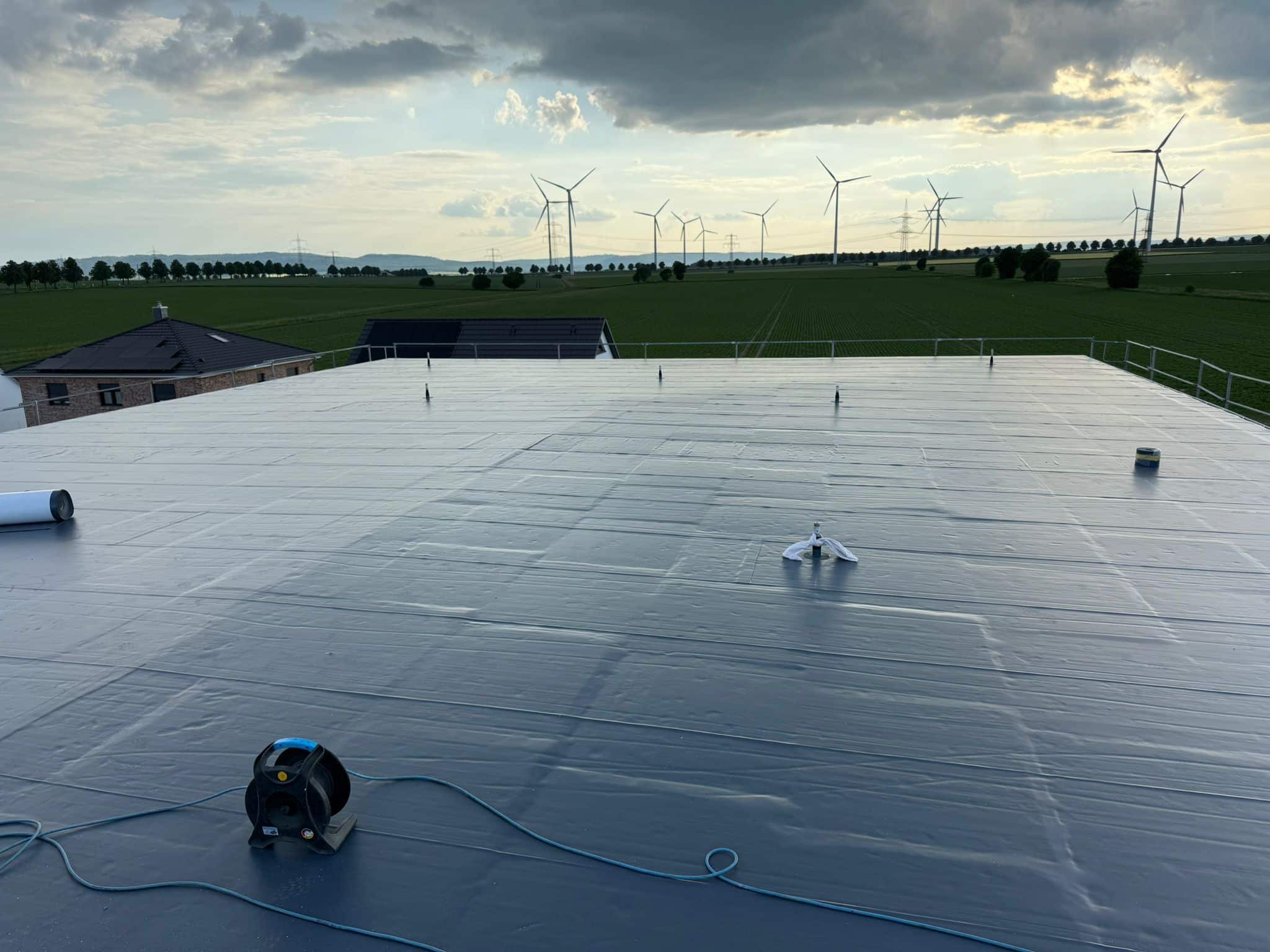 Ein Flachdach ist mit einer glatten, grauen, wasserdichten Membran bedeckt. Verschiedene Werkzeuge und Geräte, darunter eine Schlauchtrommel, liegen verstreut auf der Oberfläche. Im Hintergrund sind Windräder und Felder unter einem bewölkten Himmel zu sehen.