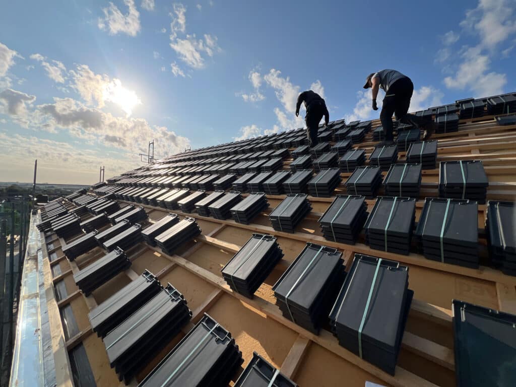 Zwei Arbeiter verlegen unter blauem Himmel mit vereinzelten Wolken dunkle Dachziegel auf einem schrägen Holzdachstuhl; das Sonnenlicht wirft Schatten auf die Ziegel.