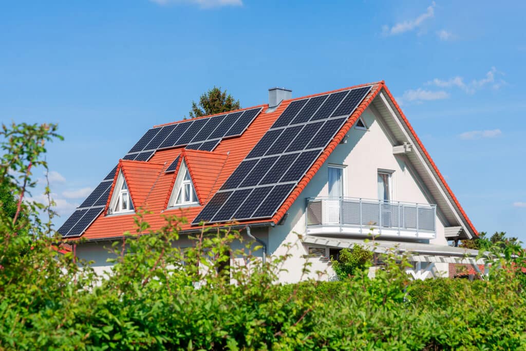 Ein modernes Haus mit einem roten Ziegeldach ist mit mehreren Sonnenkollektoren ausgestattet. Das Haus hat weiße Wände, mehrere Fenster, einen Balkon und ist umgeben von Grün unter einem blauen Himmel mit einigen Wolken.